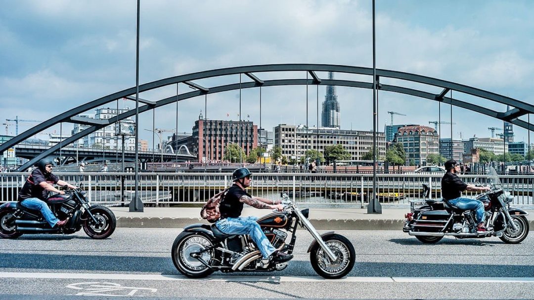 a trifecta of riders wearing military-style helmets and scooting down a bridge on a blue skied, sunny day. 