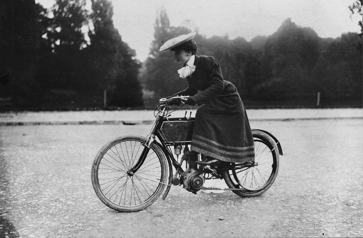 a side profile of a women trying out a motor-bicycle, popular at the turn of the century