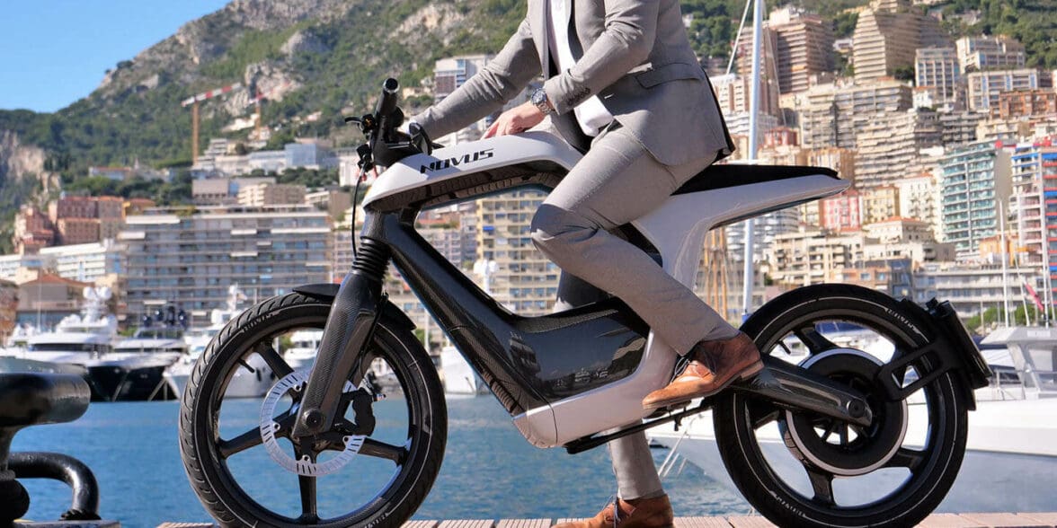 A model in a tan suit, at a dock, trying out the Novus One Electric Motorcycle with a lake in the background
