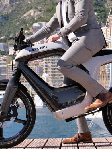 A model in a tan suit, at a dock, trying out the Novus One Electric Motorcycle with a lake in the background