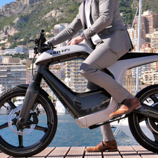 A model in a tan suit, at a dock, trying out the Novus One Electric Motorcycle with a lake in the background