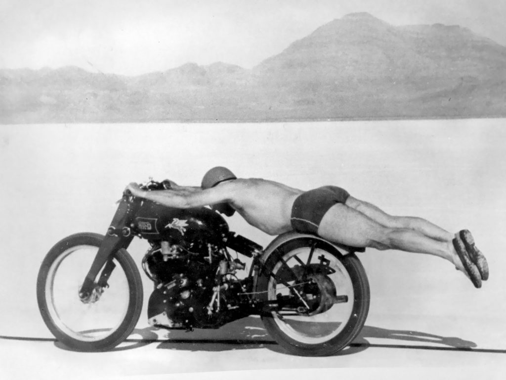 Rollie Free lying flat on rear fender of Vincent Black Shadow in speedos and swimming cap in 1950s
