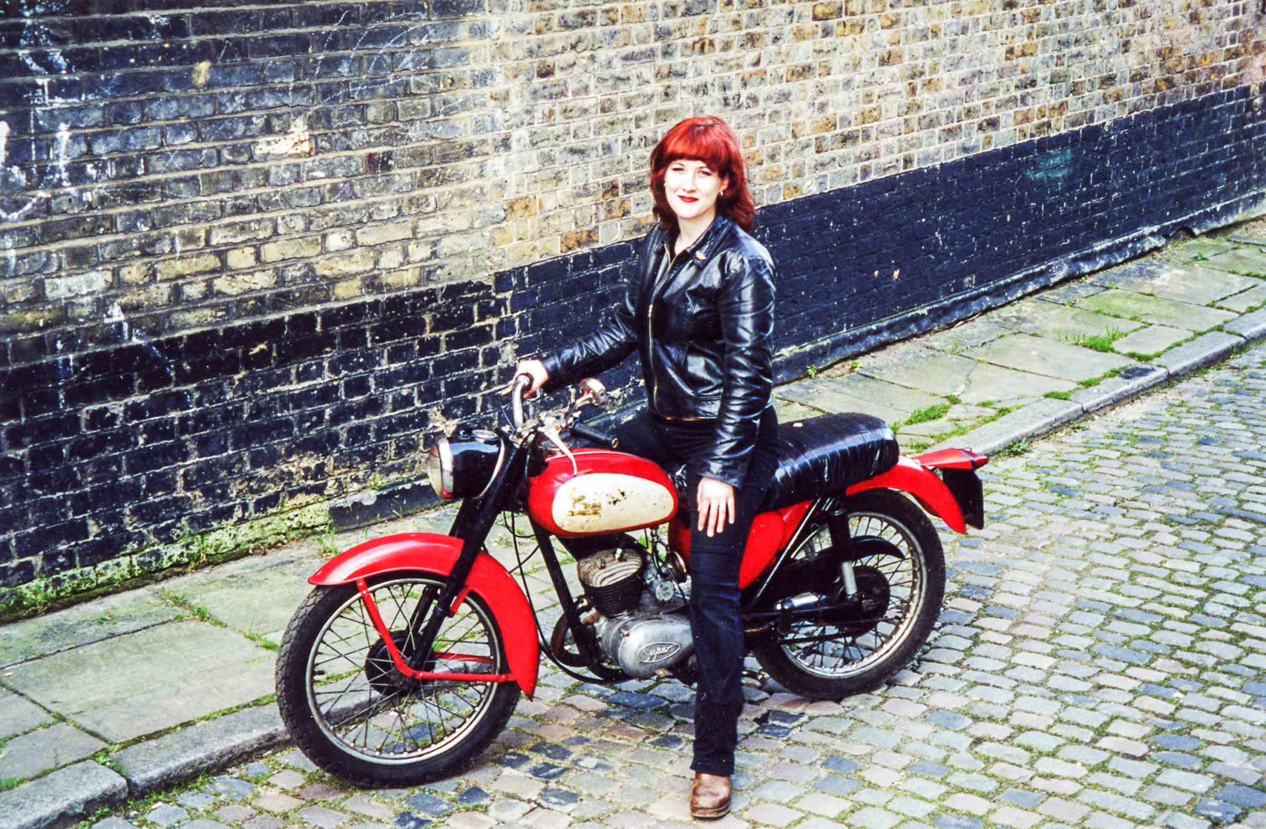 Lois Pryce astride red motorcycle on city street