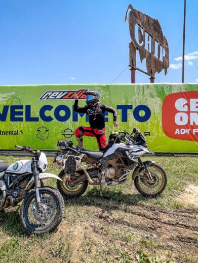 Rider posing on dirt bike in front of banner for GET ON Adv Fest by RevZilla