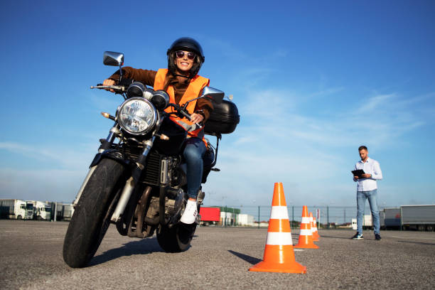 Female student with helmet taking motorcycle lessons and practicing ride. In background traffic cones and instructor with checklist rating and evaluating the ride. Motorcycle school of driving.
