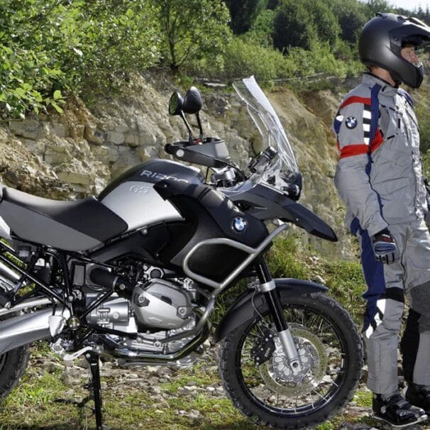 A Beemer dismounted from his BMW and standing in front of a wall of green foliage. Media sourced from Beach Moto.