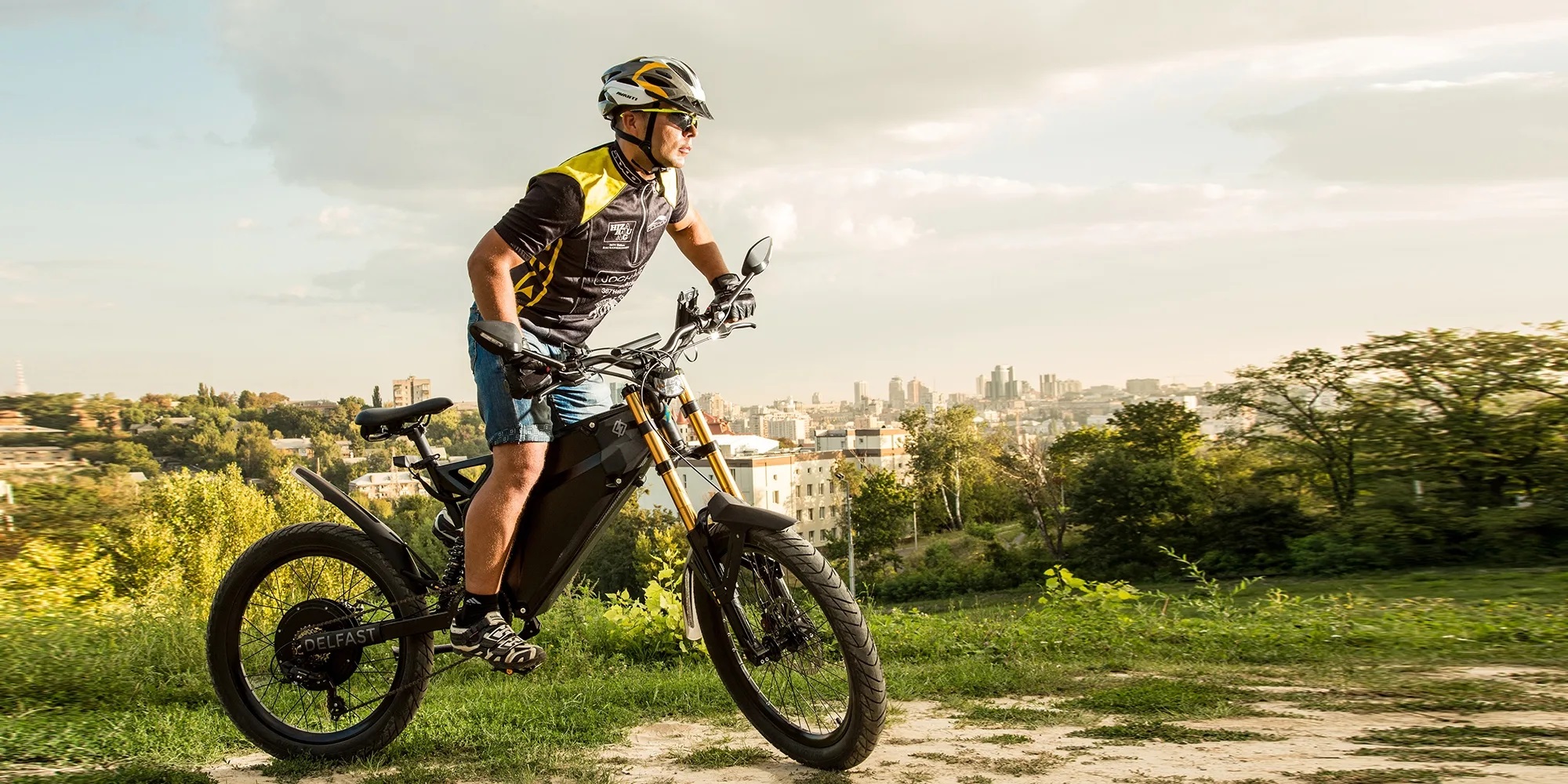 Experienced rider mounts Delfast Top 3.0 electric bicycle on grass with a large city in the backdrop