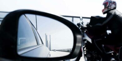 A car mirror with a motorcyclist int he background. Media sourced from RideApart.