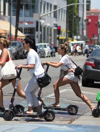Four women cross a city street with several cars in the background on kickback scooters.