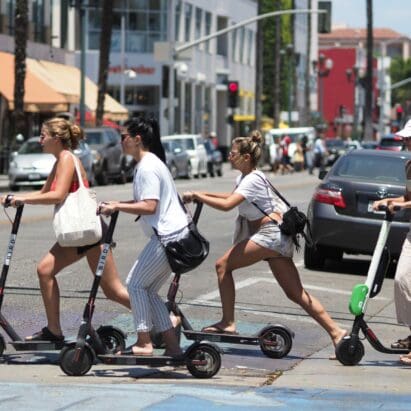 Four women cross a city street with several cars in the background on kickback scooters.