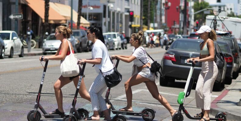 Four women cross a city street with several cars in the background on kickback scooters.