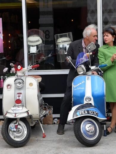An old Italian couple next to a lineup of classic Vespa scoots. Media sourced from The Guardian.