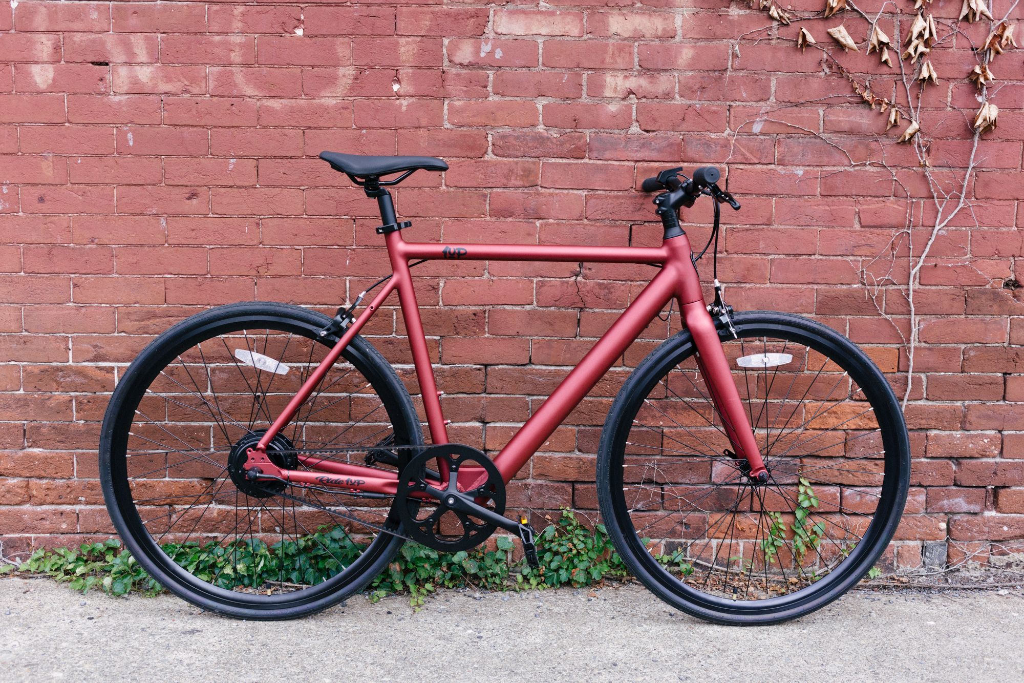 Burgundy Ride1Up ROADSTER V2 electric road bike rests against red brick wall in urban area