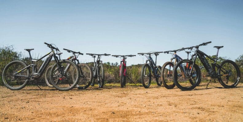 Collection of multiple electric mountain bike models lined up outside on rough terrain