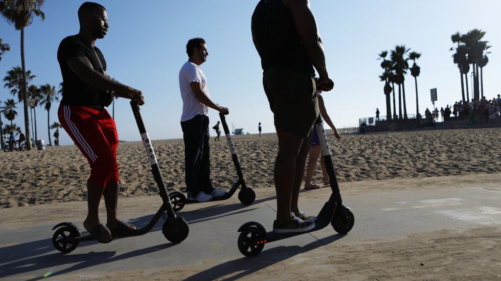 Group of friends ride Bird rental scooters down pathway on beach