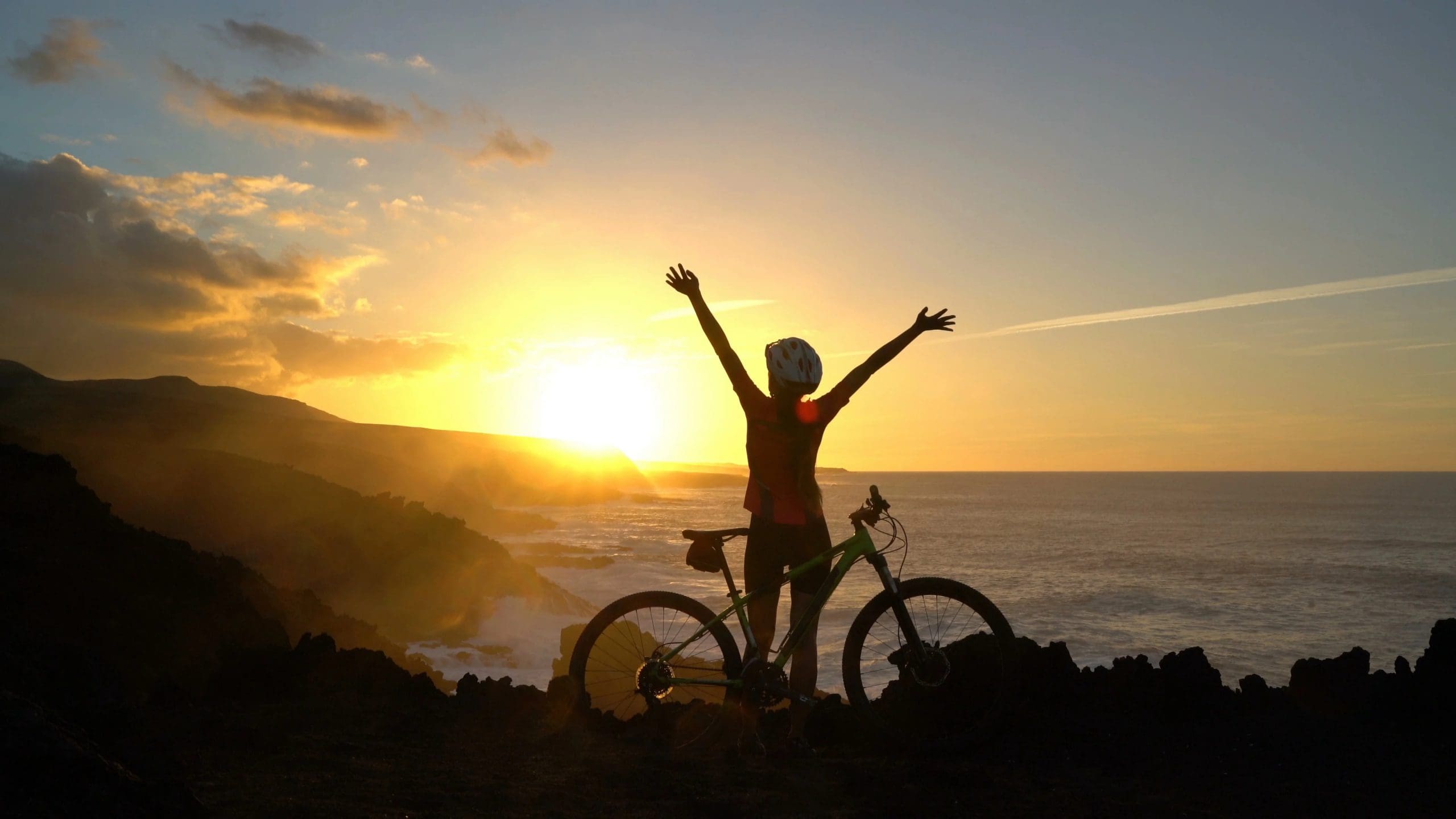 Female mountain biker on cliffs near shore with arms lifted in celebration during sunset