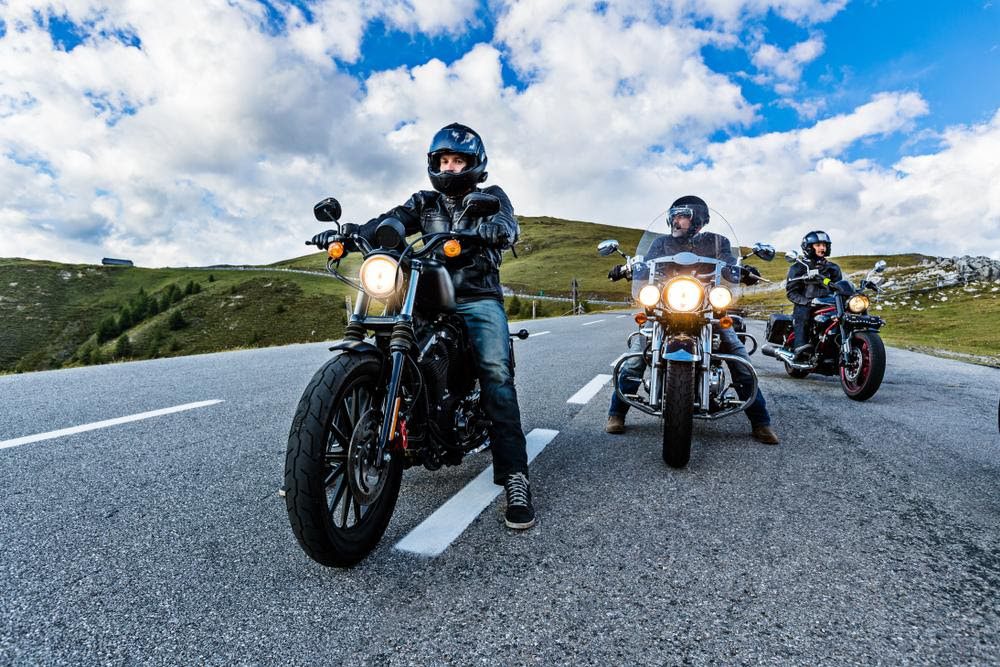 Three motorcyclists with their headlights on prepare to ride on a country road