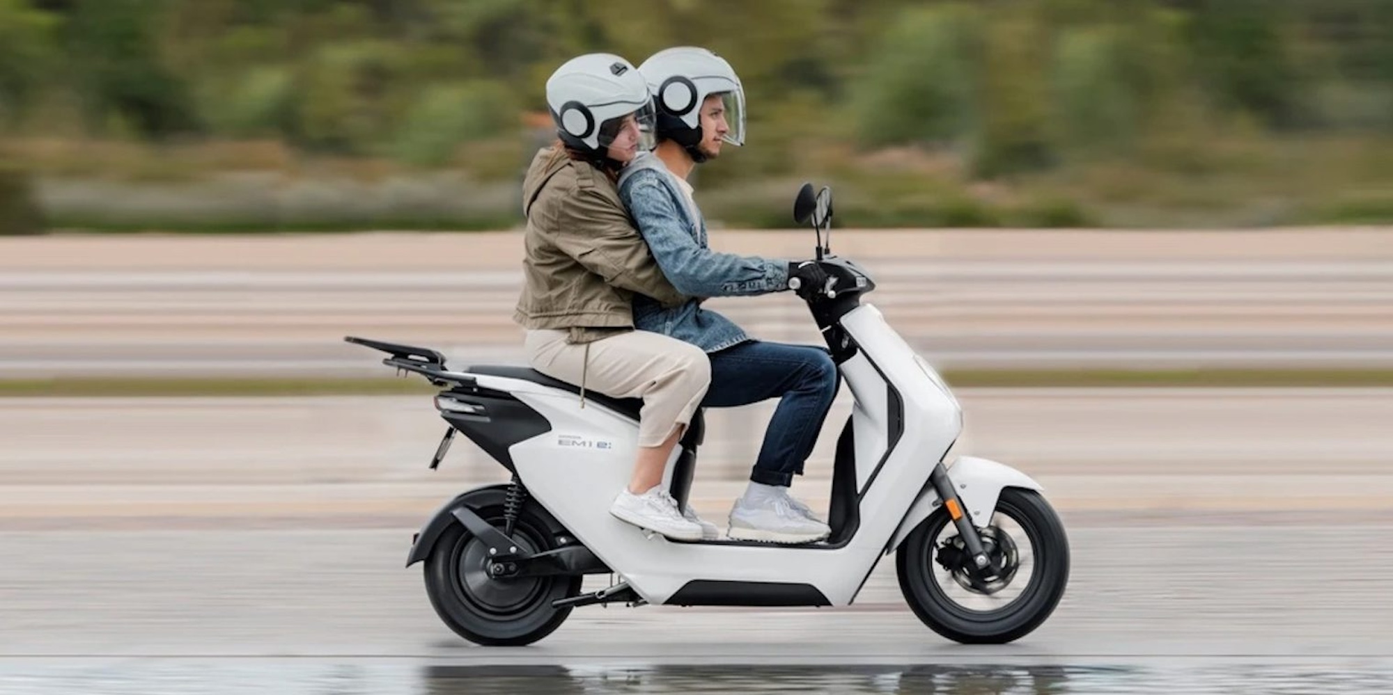 A side view of two people riding a Honda EM 1 scooter/motorcycle.